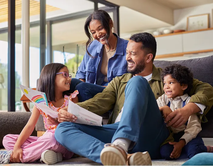 A happy family of four sitting together on the floor of a modern living room, with a young girl showing her parents a colorful drawing.