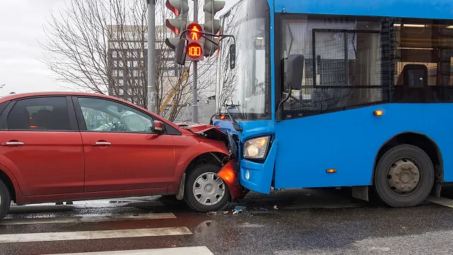 A side view of a traffic accident on a city street showing a red sedan with a crumpled front end crashed head-on into a large blue transit bus.
