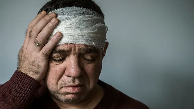 A close-up portrait of a man looking down with a grimace of pain, holding his hand to his head, which is wrapped in a thick white gauze bandage.