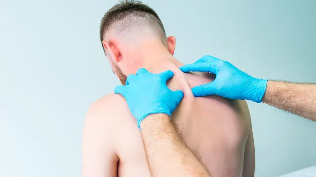 A medical practitioner wearing blue gloves examines or palpates the upper back and neck area of a shirtless male patient.