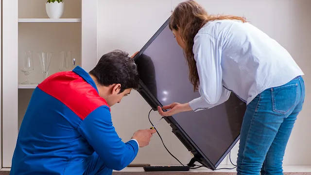 A technician in a blue and red uniform and a woman working together to lift, adjust, or install a large flat-screen television.