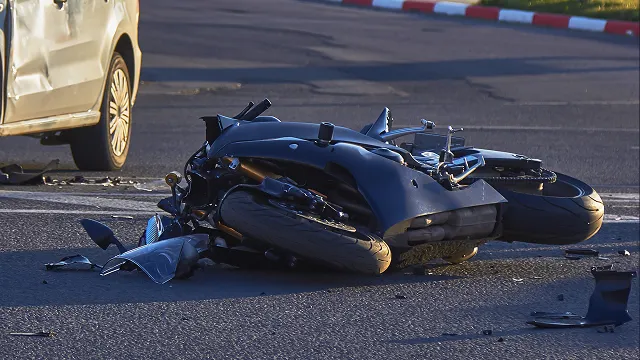 A damaged motorcycle lying on its side on an asphalt road following a traffic accident, with debris scattered nearby and part of a car visible in the background.