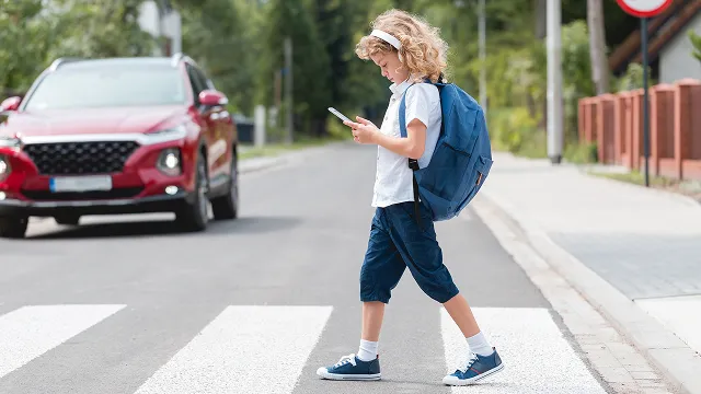A young child wearing a backpack and headphones looks down at a smartphone while walking across a pedestrian crosswalk, unaware of a red SUV approaching in the background.