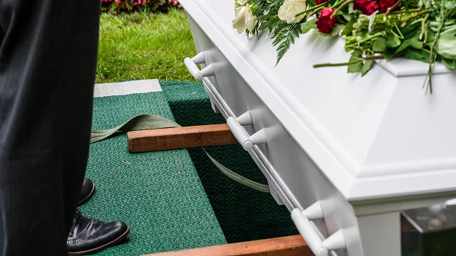 A close-up view of a white casket adorned with red and white flowers being lowered into a grave using straps at a burial service.