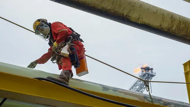 An offshore industrial worker in a red jumpsuit and safety harness balances on a beam high above the deck, with a flare boom burning in the background.