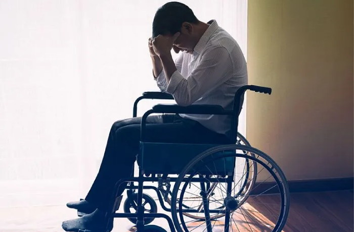A man in a light-colored button-down shirt sits in a wheelchair with his head bowed and hands pressed to his face in a gesture of distress or exhaustion. He is positioned near a bright window that casts long shadows across the wooden floor.