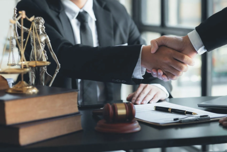 Two individuals in business attire are shaking hands over a desk, signifying a deal, settlement, or the beginning of a client-attorney relationship.